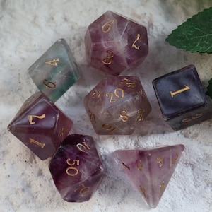 May include: A set of seven gemstone dice in different shapes, including a d4, d6, d8, d10, d12, d20, and a d100. The dice are mainly purple and clear with gold numbering. The dice are arranged on a textured, off-white surface.