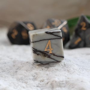 May include: A set of marbled dice with gold numbering. The focus is on a six-sided die with the number 4 in gold. Other dice in the background include a 20-sided die and a four-sided die. The dice are light gray with dark gray veining.