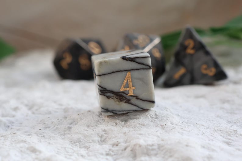 May include: A set of marbled dice with gold numbering. The focus is on a six-sided die with the number 4 in gold. Other dice in the background include a 20-sided die and a four-sided die. The dice are light gray with dark gray veining.
