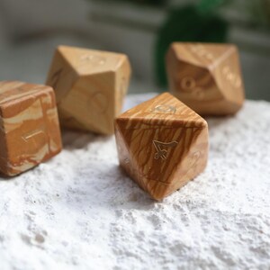 May include: A set of four polyhedral dice crafted from light brown and tan swirled material. Each die features engraved numbers with a metallic finish. The dice have various geometric shapes, including a cube and triangular-faced forms.