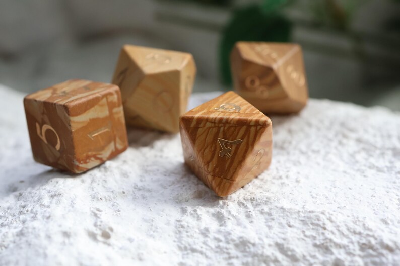 May include: A set of four polyhedral dice crafted from light brown and tan swirled material. Each die features engraved numbers with a metallic finish. The dice have various geometric shapes, including a cube and triangular-faced forms.