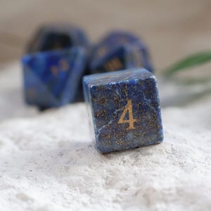 Op de afbeelding: Een vierzijdige dobbelsteen gemaakt van diepblauwe lapis lazuli steen met goudkleurige spikkels. Het cijfer vier is in een goudkleurig lettertype op de voorzijde gegraveerd. Andere blauwe stenen dobbelstenen zijn zichtbaar op de achtergrond.