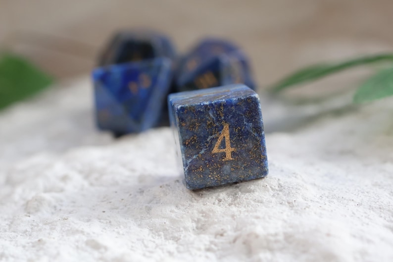 Op de afbeelding: Een vierzijdige dobbelsteen gemaakt van diepblauwe lapis lazuli steen met goudkleurige spikkels. Het cijfer vier is in een goudkleurig lettertype op de voorzijde gegraveerd. Andere blauwe stenen dobbelstenen zijn zichtbaar op de achtergrond.