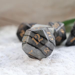 May include: A set of gray and black gemstone dice with gold numbering. The dice are in various shapes, including a 12-sided die and a triangular die. The dice are set on a white, textured surface.