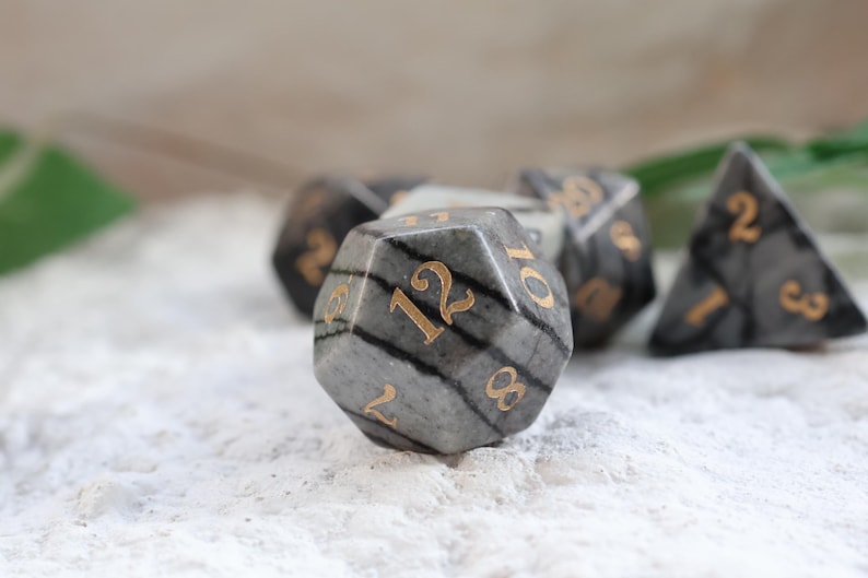 May include: A set of gray and black gemstone dice with gold numbering. The dice are in various shapes, including a 12-sided die and a triangular die. The dice are set on a white, textured surface.