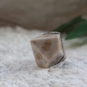 May include: A multi-sided, beige-colored die with gold-colored numbers. The die has a marbled appearance and is marked with the numbers 30 and 40. A black band wraps around the die. The die is resting on a textured, white surface.