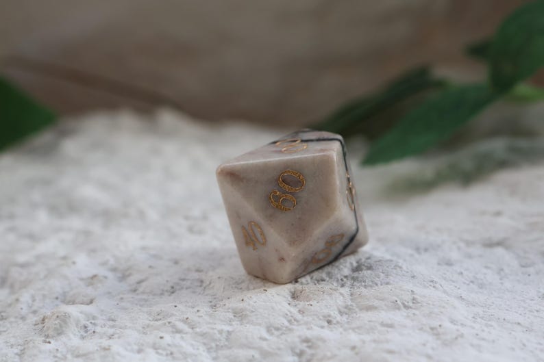 May include: A multi-sided, beige-colored die with gold-colored numbers. The die has a marbled appearance and is marked with the numbers 30 and 40. A black band wraps around the die. The die is resting on a textured, white surface.