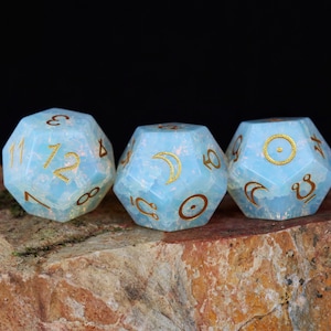 May include: Three light blue, twelve-sided dice with gold-colored numbers and symbols. The dice are set against a dark background and rest on a textured, brown and orange stone surface. The dice are for tabletop games.