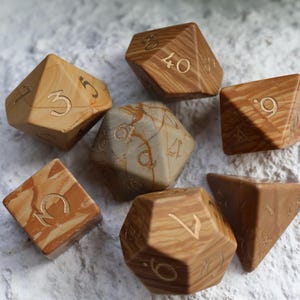 May include: A set of seven polyhedral dice featuring a tan and brown marbled pattern with engraved numbers. The collection includes various geometric shapes such as a cube, a tetrahedron, and icosahedrons, all displaying a smooth, polished finish.
