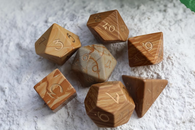 May include: A set of seven polyhedral dice featuring a tan and brown marbled pattern with engraved numbers. The collection includes various geometric shapes such as a cube, a tetrahedron, and icosahedrons, all displaying a smooth, polished finish.