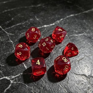 Puede incluir: Un juego de siete dados poliédricos de color rojo translúcido con números dorados. La colección incluye un d20, d12, d10, d8, d6, d4 y un dado de porcentaje, todos con bordes afilados y un acabado pulido que captura la luz.