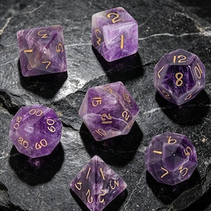 May include: A set of seven polyhedral dice carved from purple amethyst gemstone with engraved gold-coloured numbers. The collection includes various geometric shapes such as a d20, d12, d10, d8, d6, and d4, all featuring polished facets and sharp edges.