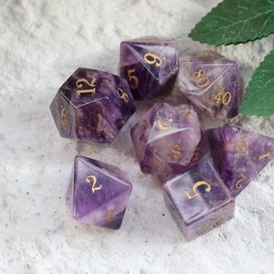 May include: Seven purple and gold dice with various numbers of sides. The dice are a translucent purple with gold numbering. They are arranged on a white, textured surface, with green leaves in the background. A set of dice for tabletop games.