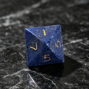 Op de afbeelding: Een vierzijdige piramidevormige dobbelsteen gemaakt van diepblauwe lapis lazuli steen met goudkleurige gegraveerde cijfers. De zichtbare zijden tonen de cijfers 1, 3, 5 en 7, die in het gepolijste oppervlak van het donkerblauwe mineraal zijn ge&euml;tst.
