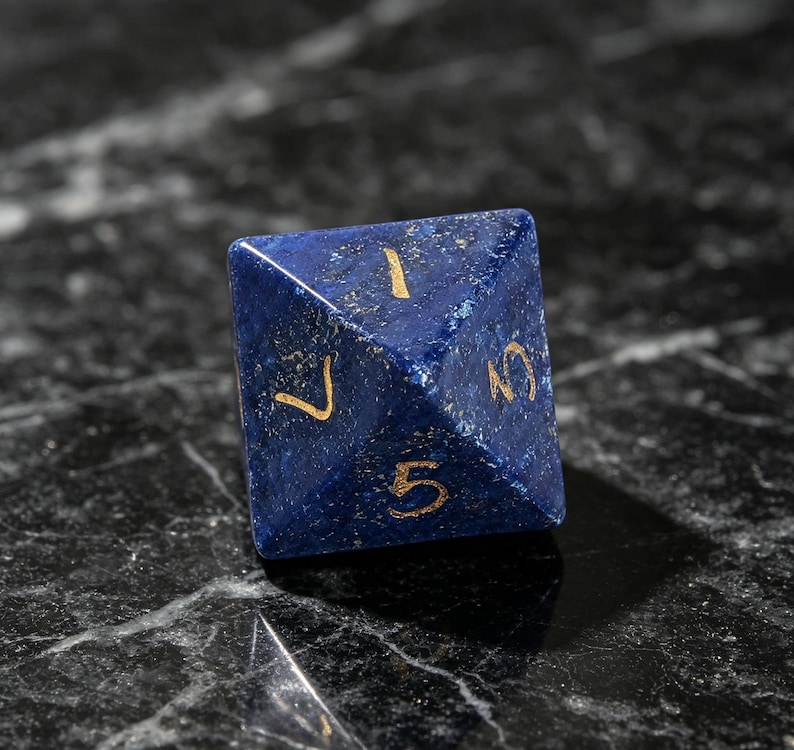 Op de afbeelding: Een vierzijdige piramidevormige dobbelsteen gemaakt van diepblauwe lapis lazuli steen met goudkleurige gegraveerde cijfers. De zichtbare zijden tonen de cijfers 1, 3, 5 en 7, die in het gepolijste oppervlak van het donkerblauwe mineraal zijn ge&euml;tst.