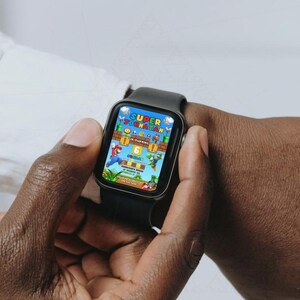 May include: A black smartwatch displaying a colorful video game scene with the text "SUPER JORNATAN". The game features a cartoon character in a red outfit. The watch is on a wrist.