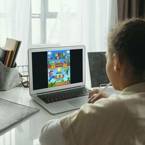 May include: A laptop displays a colorful Super Mario Bros. graphic. A person is using a stylus on the screen. A gray felt envelope and a gray organizer with books are on the white desk. The scene is lit by natural light.