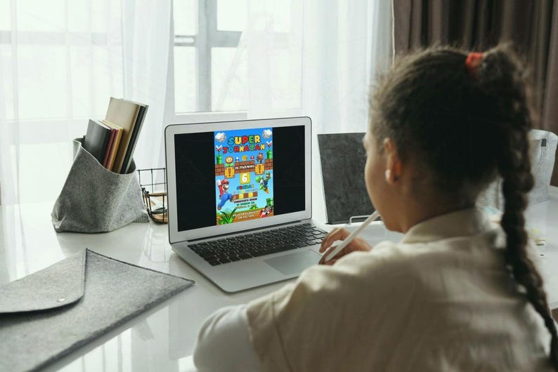 May include: A laptop displays a colorful Super Mario Bros. graphic. A person is using a stylus on the screen. A gray felt envelope and a gray organizer with books are on the white desk. The scene is lit by natural light.