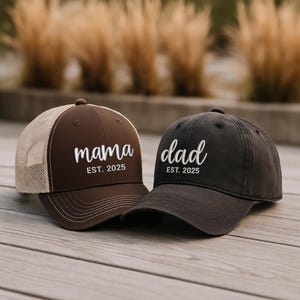 Pu&ograve; includere: Due cappellini da baseball, uno marrone con retro in rete beige e la scritta "mama" ricamata in bianco, e l'altro grigio scuro con "dad" ricamato in bianco. Entrambi i cappellini hanno "EST. 2025" sotto.