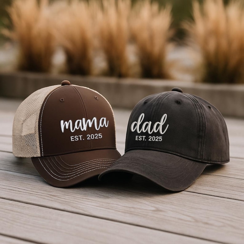 Pu&ograve; includere: Due cappellini da baseball, uno marrone con retro in rete beige e la scritta "mama" ricamata in bianco, e l'altro grigio scuro con "dad" ricamato in bianco. Entrambi i cappellini hanno "EST. 2025" sotto.