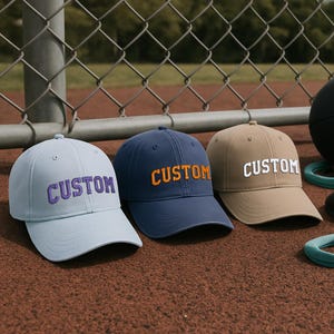 May include: Three baseball caps in light blue, navy, and tan, each embroidered with the word "CUSTOM" in a contrasting color. The caps are displayed on a textured surface, with a chain-link fence in the background.