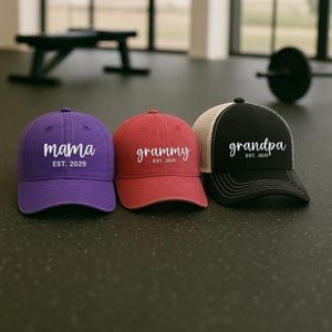 Pu&ograve; includere: Tre cappellini da baseball viola, rossi e neri. Il cappellino viola dice "mama" con "EST. 2025" sotto. Il cappellino rosso dice "grammy" con "EST. 2025". Il cappellino nero dice "grandpa" con "EST. 2025".