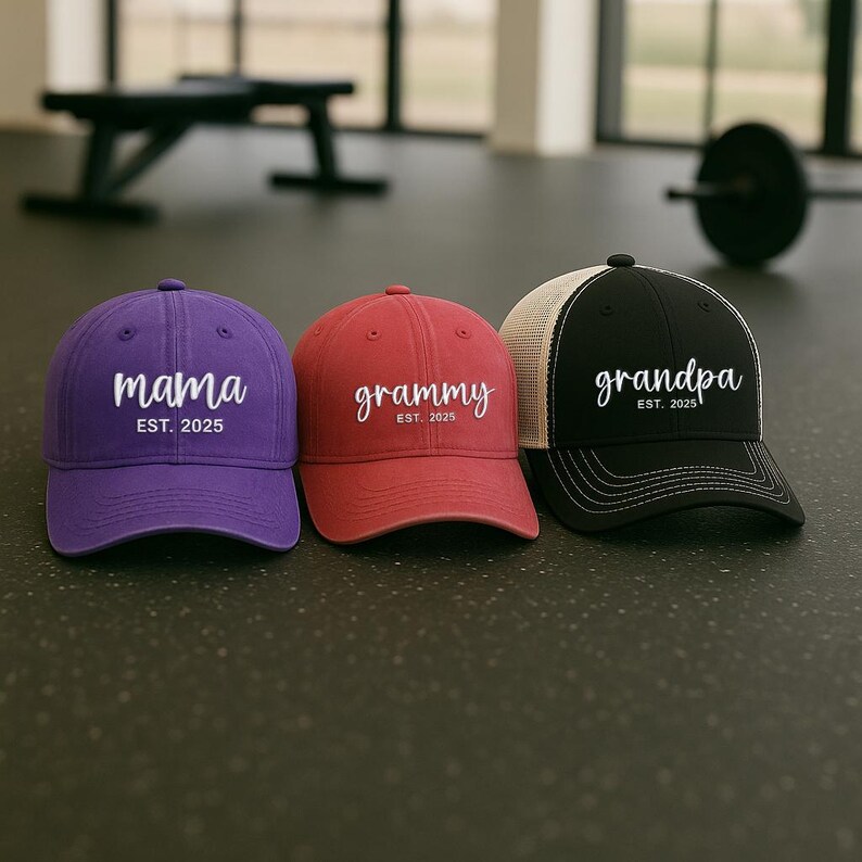 Pu&ograve; includere: Tre cappellini da baseball viola, rossi e neri. Il cappellino viola dice "mama" con "EST. 2025" sotto. Il cappellino rosso dice "grammy" con "EST. 2025". Il cappellino nero dice "grandpa" con "EST. 2025".