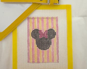 May include: Yellow luggage tag with a clear window and a stitched canvas design. The canvas features a black Minnie Mouse head with a pink bow, set against a background of pink and yellow stripes. The tag has a yellow strap with a silver buckle.