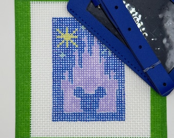 May include: A needlepoint canvas featuring a light purple castle silhouette with a Mickey Mouse head shape at the base, set against a dark blue background with a yellow star. A bright blue leather luggage tag strap rests across the top corner.