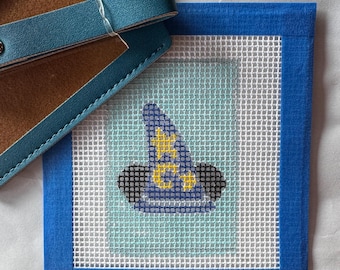 May include: A blue needlepoint coaster featuring a wizard hat design in blue, yellow, and black. The coaster is framed with a blue border and sits next to a blue leather luggage tag.
