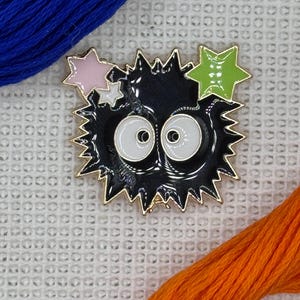 May include: A black enamel pin shaped like a spiky, round creature with two large white eyes. The pin features a small pink star and a larger lime green star attached to the top. The design is outlined in gold metal and has a glossy finish.