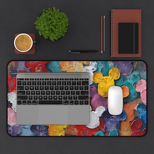 May include: A desk setup featuring a laptop, mouse, and a colorful mousepad with a Mickey Mouse design. The mousepad has a vibrant, abstract painted background with yellow, blue, purple, and red hues. A cup of coffee, a plant, and stationery are also present.