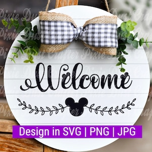 Puede incluir: Un letrero redondo de madera blanco con un lazo a cuadros blanco y negro, hojas verdes y la palabra "Bienvenido" en letra negra. Una silueta negra de la cabeza de Mickey Mouse se encuentra debajo de la palabra "Bienvenido" con una guirnalda de hojas negras.