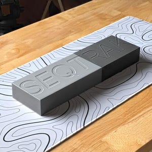 May include: A rectangular, two-toned gray object with the word "SEQTRAK" embossed on its surface. The object is resting on a white and black topographical map-patterned mat, placed on a wooden surface.