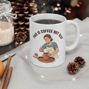 May include: White ceramic mug with the text "THIS IS COFFEE NOT SLIP" and an illustration of a person working with clay. The mug is filled with coffee and sits on a light-colored surface, with decorative elements.