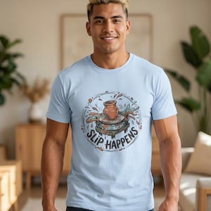 May include: A light blue t-shirt featuring a pottery wheel design with the text "Slip Happens" in a circular pattern. The design includes a brown clay pot and splashes of color. The shirt is worn by a person.
