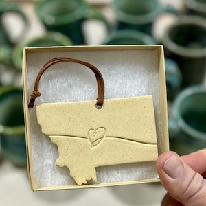 May include: A ceramic ornament in the shape of the state of Montana, with a heart design. The ornament is a light beige color and has a brown leather cord for hanging. It is presented in a small cardboard box.