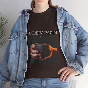 Puede incluir: Camiseta marrón oscuro con el texto "MUDDY POTS" en blanco. La camiseta presenta una imagen de manos sosteniendo una olla de cerámica oscura. La persona lleva una chaqueta vaquera azul y vaqueros azules.