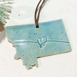 May include: A ceramic ornament in the shape of the state of Montana, with a light blue glaze and a brown leather cord for hanging. The ornament features a heart design. The ornament is speckled with dark spots.