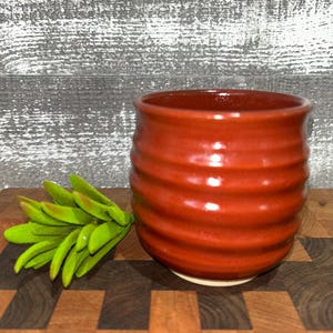 May include: A handmade, terracotta-colored ceramic cup with a ribbed exterior. The cup sits on a wooden surface, with a sprig of green leaves to the left. The cup has a smooth, glossy finish and a white base.