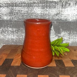 May include: A handmade, red ceramic vase with a speckled texture. The vase has a wide base that tapers inward and then flares out at the top. The vase is displayed on a wooden cutting board with a green plant sprig.