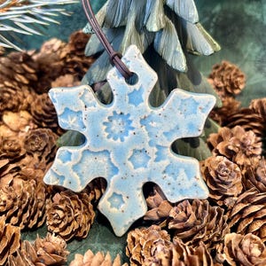 May include: A light blue ceramic snowflake ornament with a speckled finish and embossed star patterns. The ornament features a hole at the top and is suspended by a thin brown leather cord, showcasing a glossy glaze and six-pointed design.
