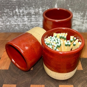 May include: Three handmade ceramic match holders in a rustic style. The holders are glazed in a rich red color, with a speckled beige base on one. One holder is filled with matches with green tips. The holders are on a wooden surface.