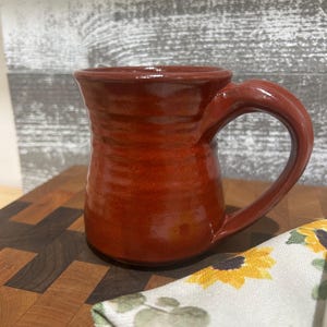 May include: A handmade, reddish-brown ceramic mug with a curved handle. The mug has a glossy finish and a textured surface. It sits on a wooden cutting board, with a floral patterned cloth in the foreground. The mug is designed for hot beverages.