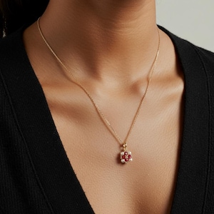 May include: A gold necklace with a pendant featuring a flower design. The pendant has a central cluster of red stones surrounded by white pearls. The necklace is worn against a black top.