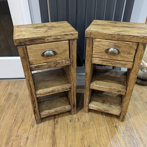 May include: Two matching bedside tables crafted from light brown wood with a visible grain. Each unit features a single top drawer with a rounded metal cup pull handle, followed by two open storage shelves below, providing a compact vertical design.
