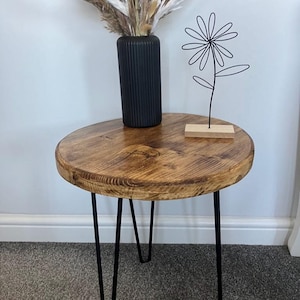 May include: A round wooden side table with a natural finish, supported by black hairpin legs. A black vase with dried decorative grasses and a wire flower sculpture sit on the table. The table's diameter is approximately 61 cm.