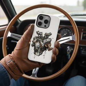 Puede incluir: Funda de teléfono blanca con una ilustración detallada de un motor. El diseño muestra pistones, tubos de escape y piezas mecánicas. La funda se sostiene con un guante de cuero marrón, con un volante de madera en el fondo.