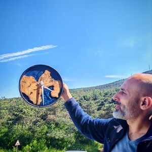 Puede incluir: Un reloj de madera redondo con un diseño de vetas de madera natural y detalles de resina azul. Las manecillas del reloj son blancas. El reloj se sostiene contra un fondo de cielo azul y árboles verdes.