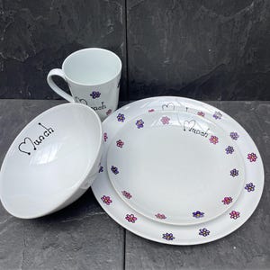 May include: A white mug, bowl, and two plates with a floral design. The mug and bowl have the word "Much" written on them. The plates have a pink and purple floral design.